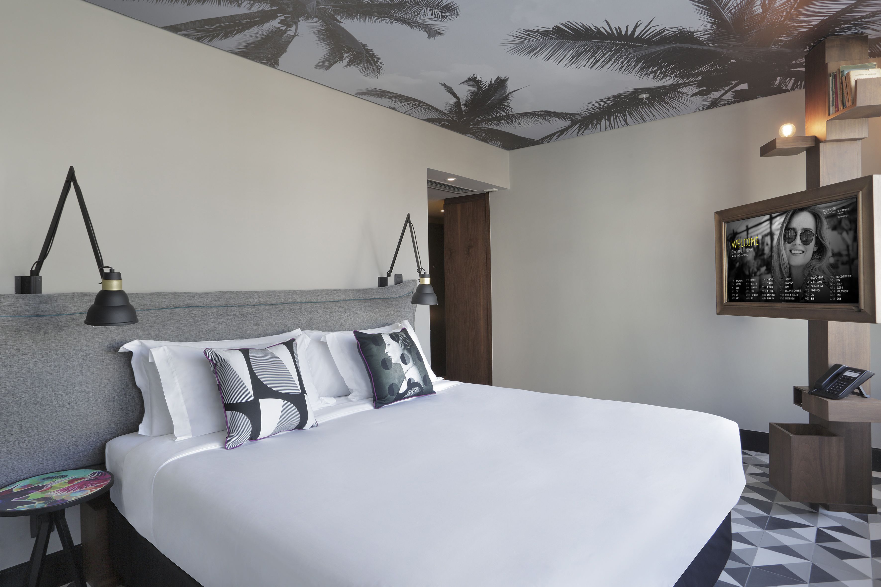 Una habitación de hotel moderna con una cama grande, una mesita de noche, una lámpara de lectura y un cartel de bienvenida enmarcado en la pared.