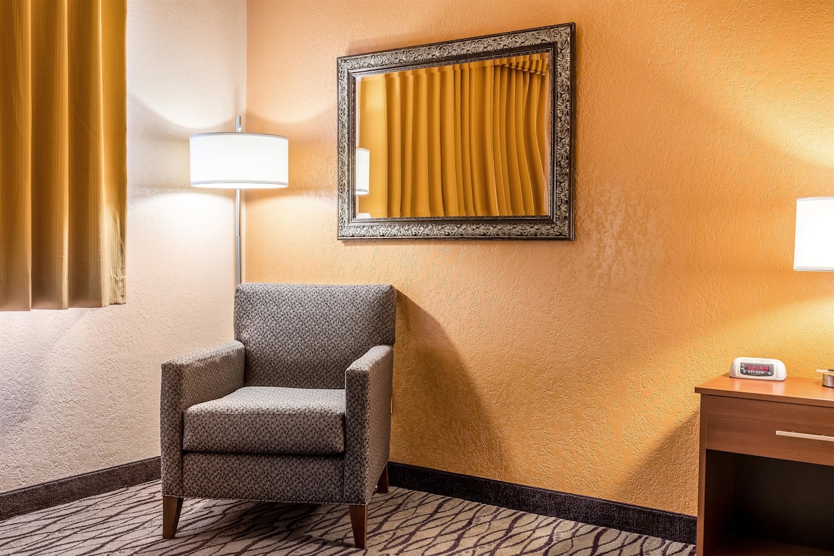 A hotel room with a patterned armchair, a large mirror, a bedside table with a lamp, and a patterned carpet.