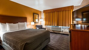 A hotel room with a large bed, a desk, a chair, a mirror, a window with curtains, and an air conditioning unit. - SureStay Hotel by Best Western Wenatchee (East Wenatchee)