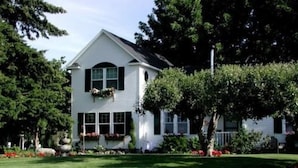 Une maison blanche à deux étages, avec un toit à pignon, un porche à l’avant orné d’une jardinière et une pelouse bien entretenue.