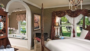 Une chambre à coucher avec un lit à baldaquin, un coin salon avec un fauteuil, une coiffeuse avec un miroir et une vue sur l’extérieur.
