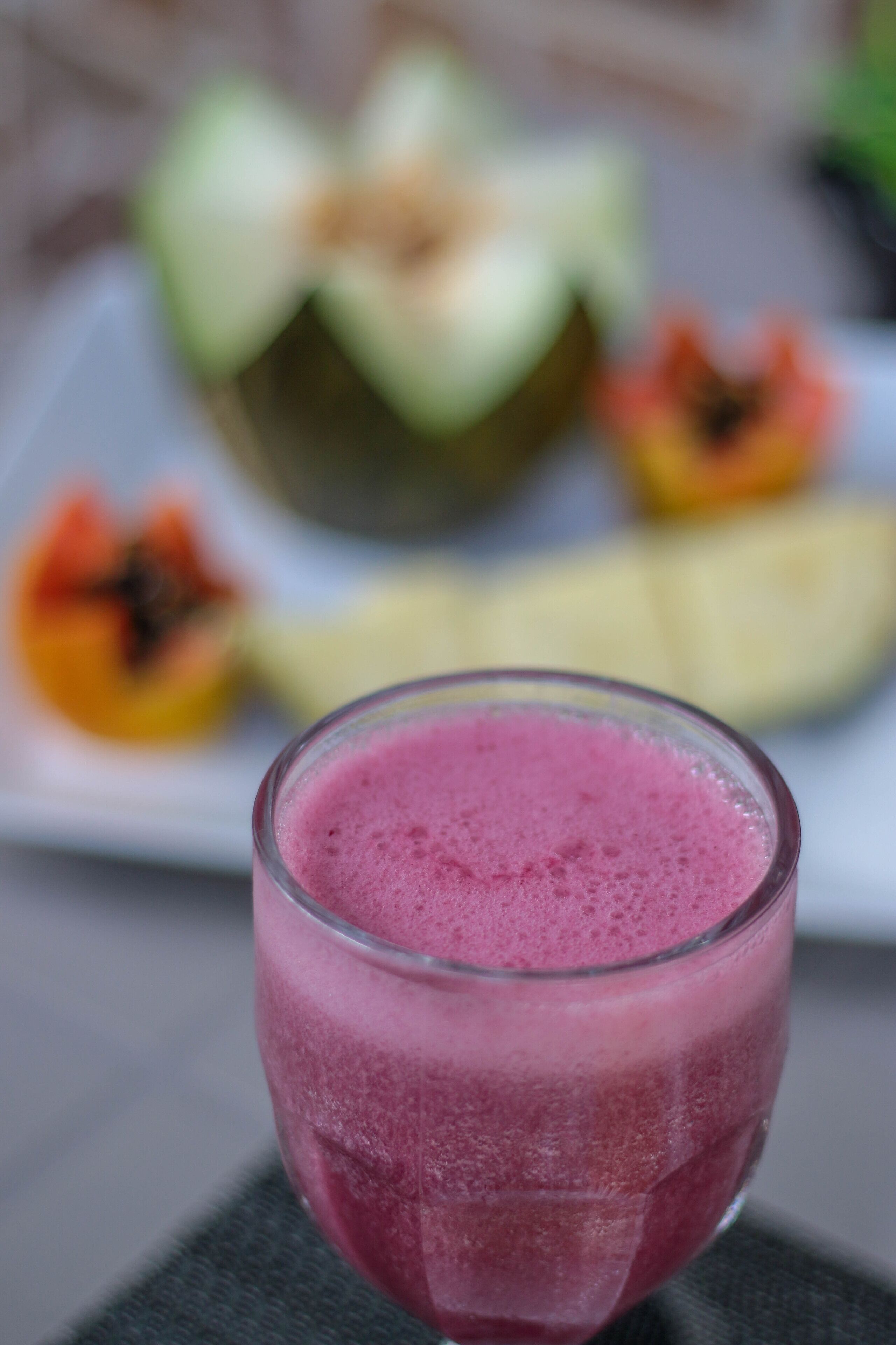 Un vaso de batido rosa sobre una mesa con un plato de frutas al fondo.