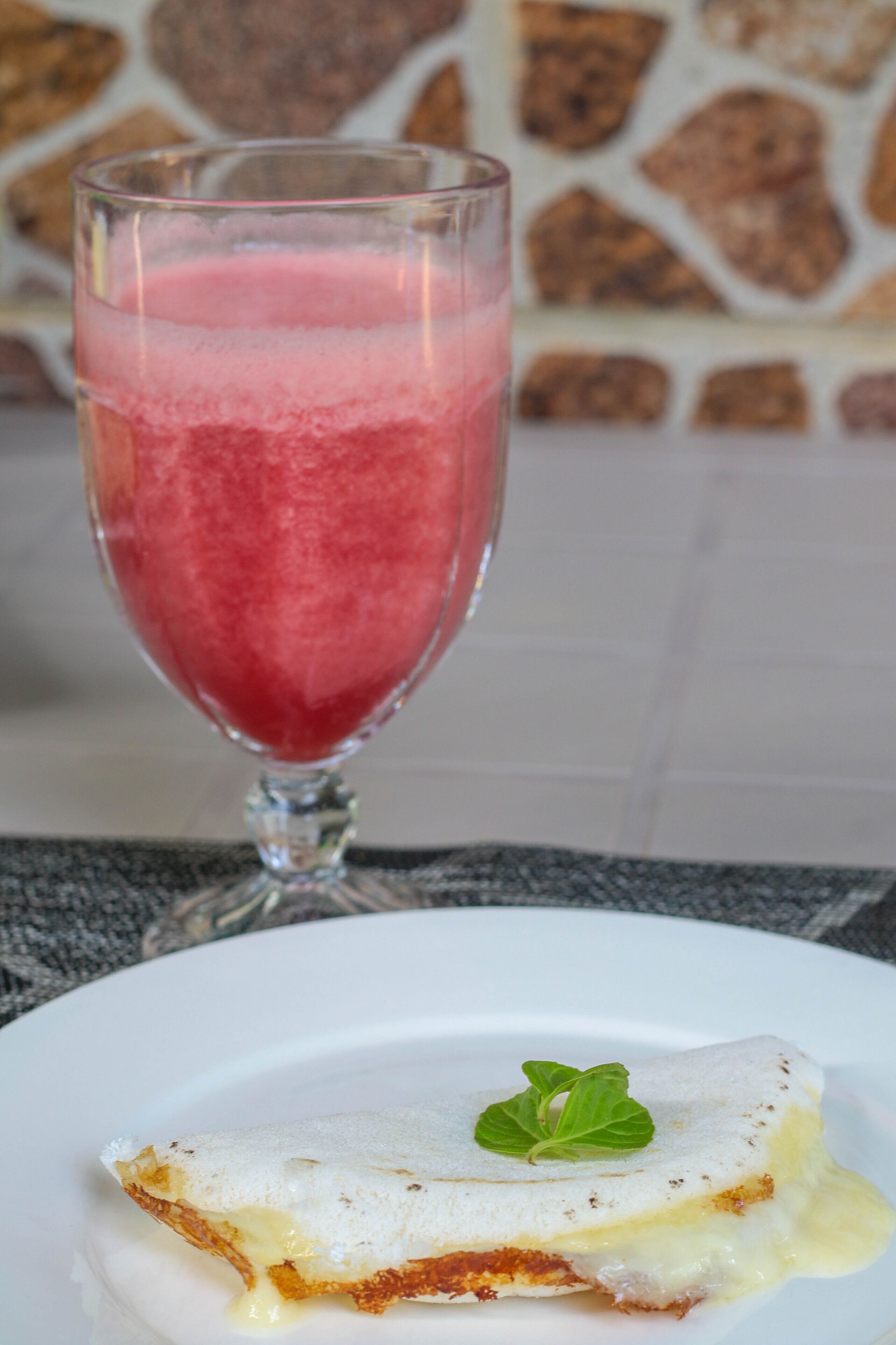 A dessert with a slice of cake topped with a mint leaf, a glass of pink beverage, and a stone wall in the background.