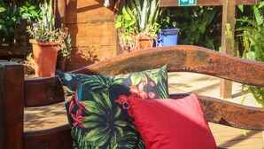 A wooden bench with floral upholstery and a red pillow, surrounded by potted plants and a wooden structure with a mirror. - Pousada Leão Marinho (Fernando de Noronha)