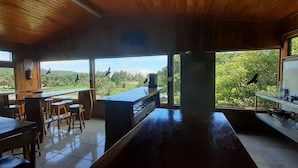 A kitchen with a large island, wooden tables, and chairs, and large windows offering a view of greenery. - Hostel La Suerte (Monteverde)