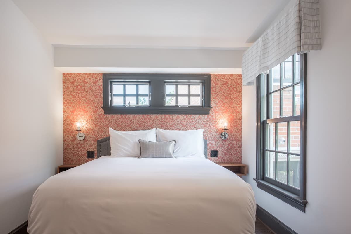 Una cama bien tendida con sábanas blancas, dos apliques de pared y una ventana con vistas a un edificio de ladrillo.