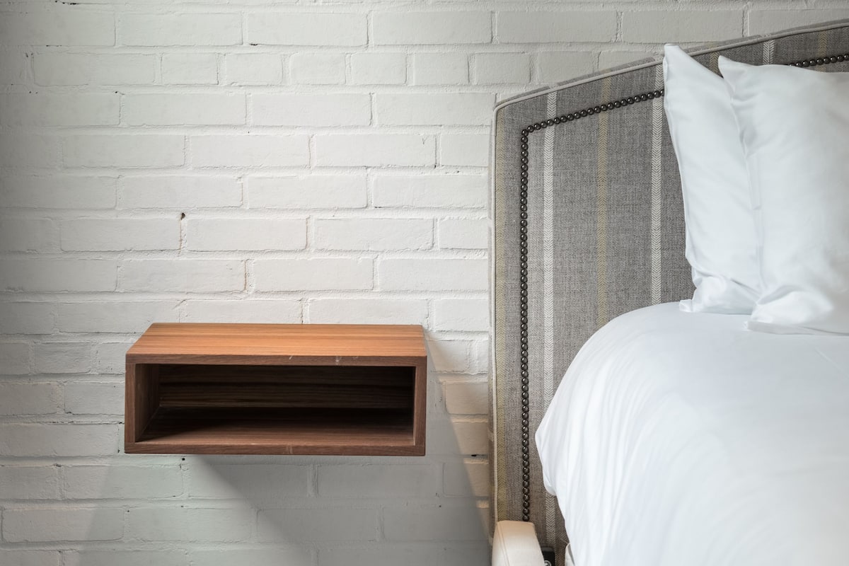 Un dormitorio con pared de ladrillo blanco, una lámpara colgante, una mesita de noche con estante de madera y una cama con ropa blanca.