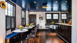 A restaurant with wooden flooring, round tables, and black chairs. There are windows with blinds and a dark ceiling. - The Poppy Georgetown (Washington)