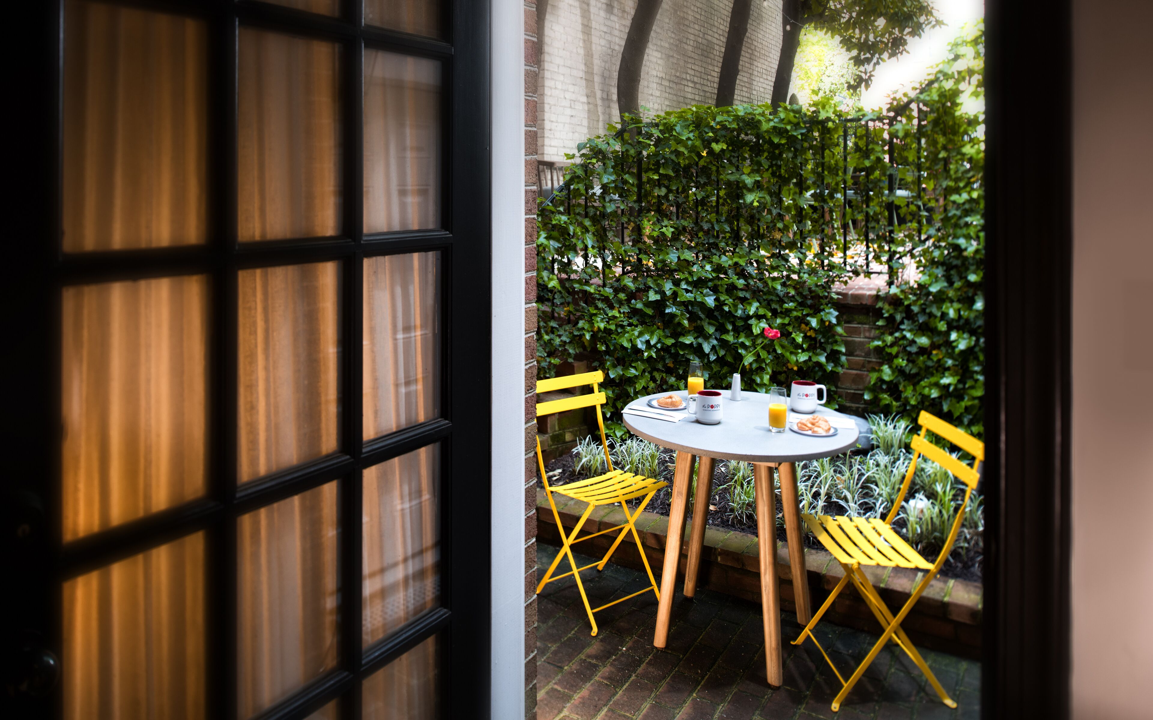 A patio with a round table and yellow chairs, surrounded by greenery and a brick wall.