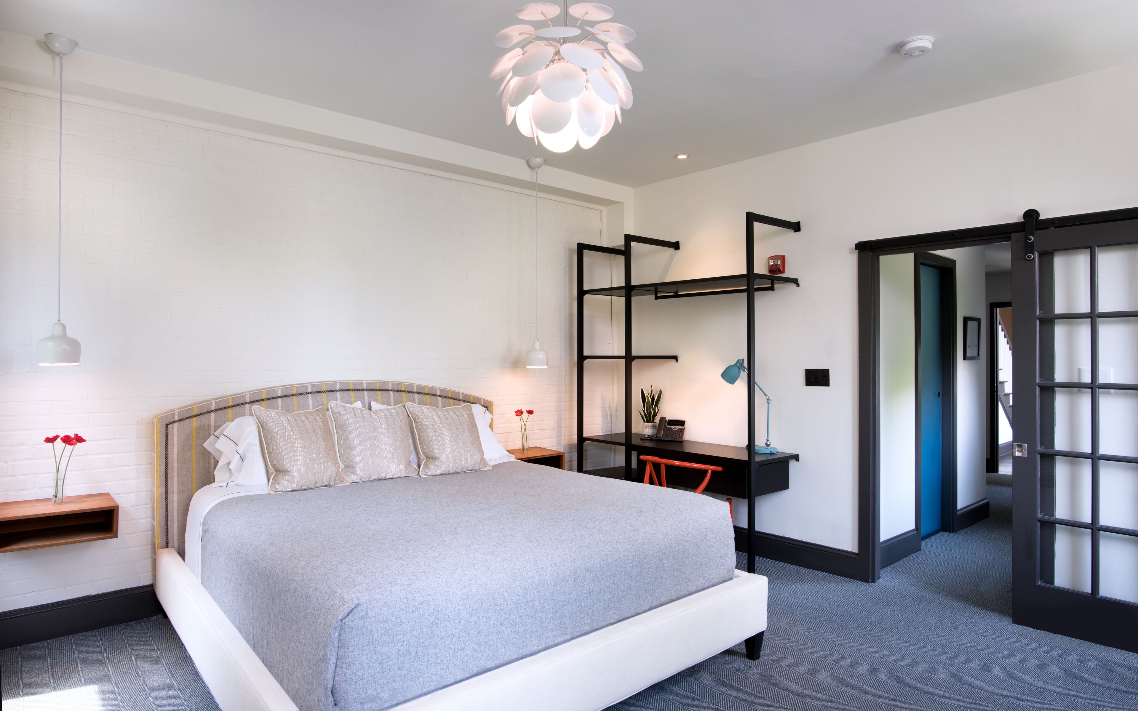 Um quarto moderno com cama, mesas de cabeceira, escrivaninha e cadeira. Há uma estante de livros, uma luminária pendente e uma porta que dá para outro cômodo.