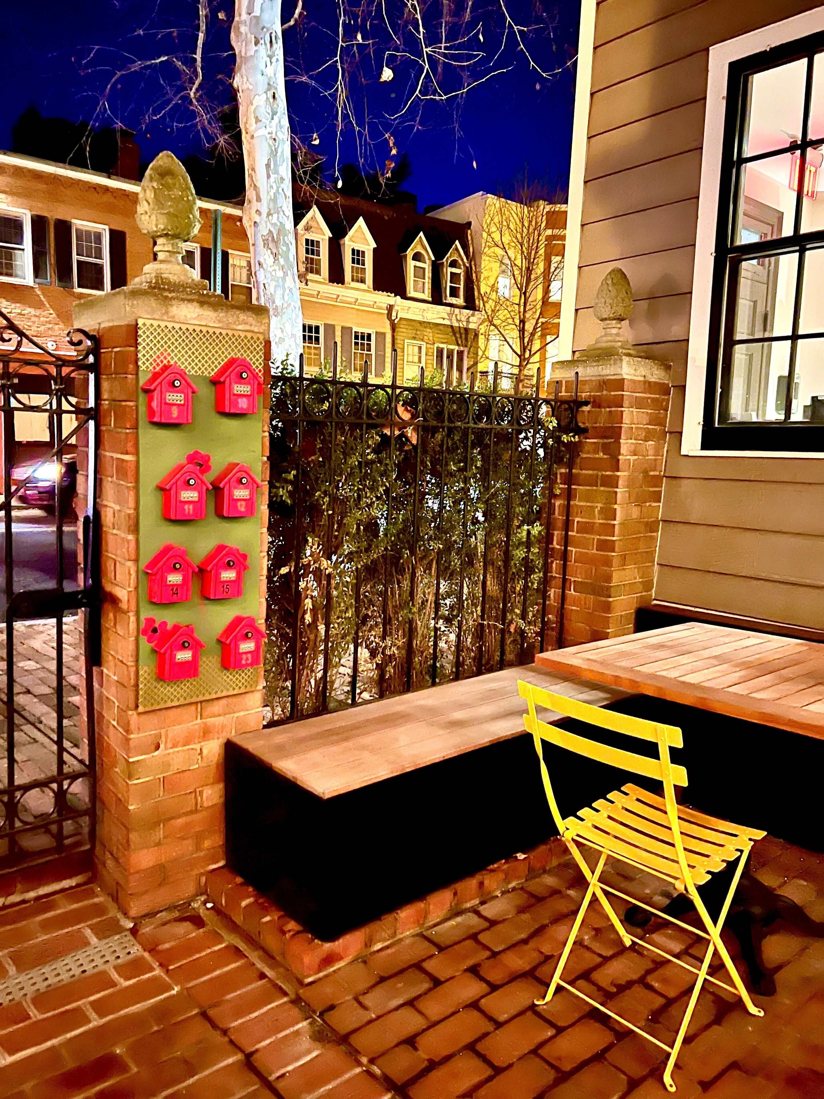 Une terrasse en briques avec une table en bois et des chaises jaunes, une clôture décorative ornée de nichoirs numérotés, et un bâtiment avec de nombreuses fenêtres en arrière-plan.