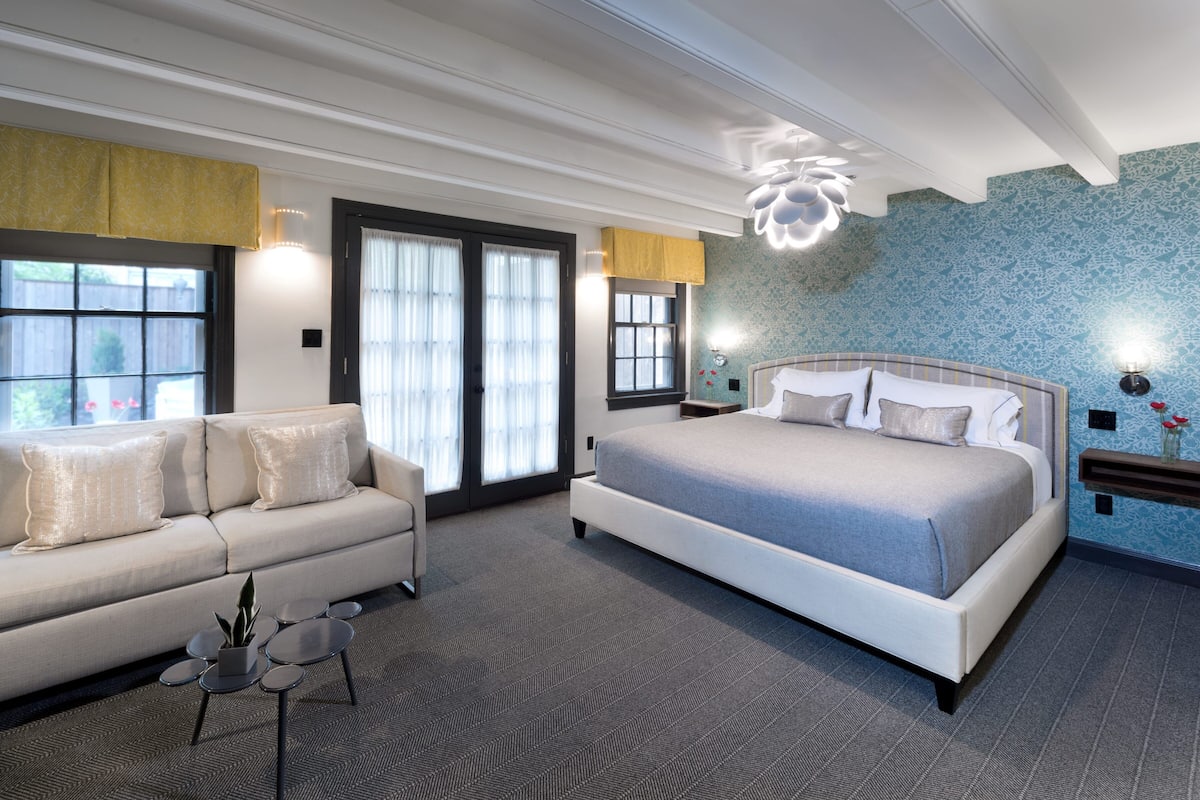 Una habitación de hotel moderna con una cama grande, un sofá, una pequeña mesa con una planta y una lámpara de pared decorativa.