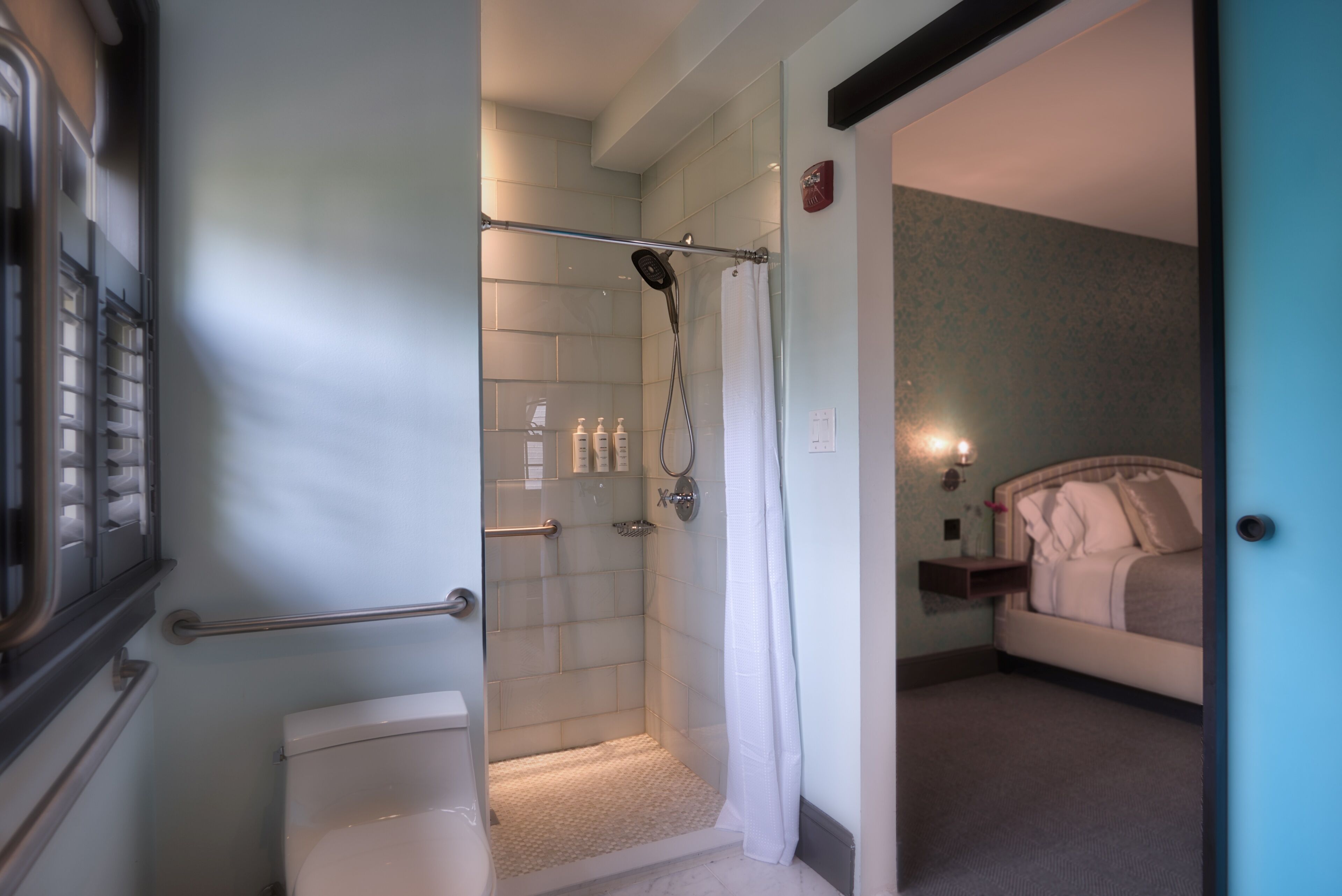 A hotel bathroom with a shower, toilet, and a bed visible in the adjacent room.
