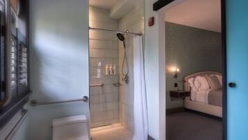 A hotel bathroom with a shower, toilet, and a bed visible in the adjacent room.