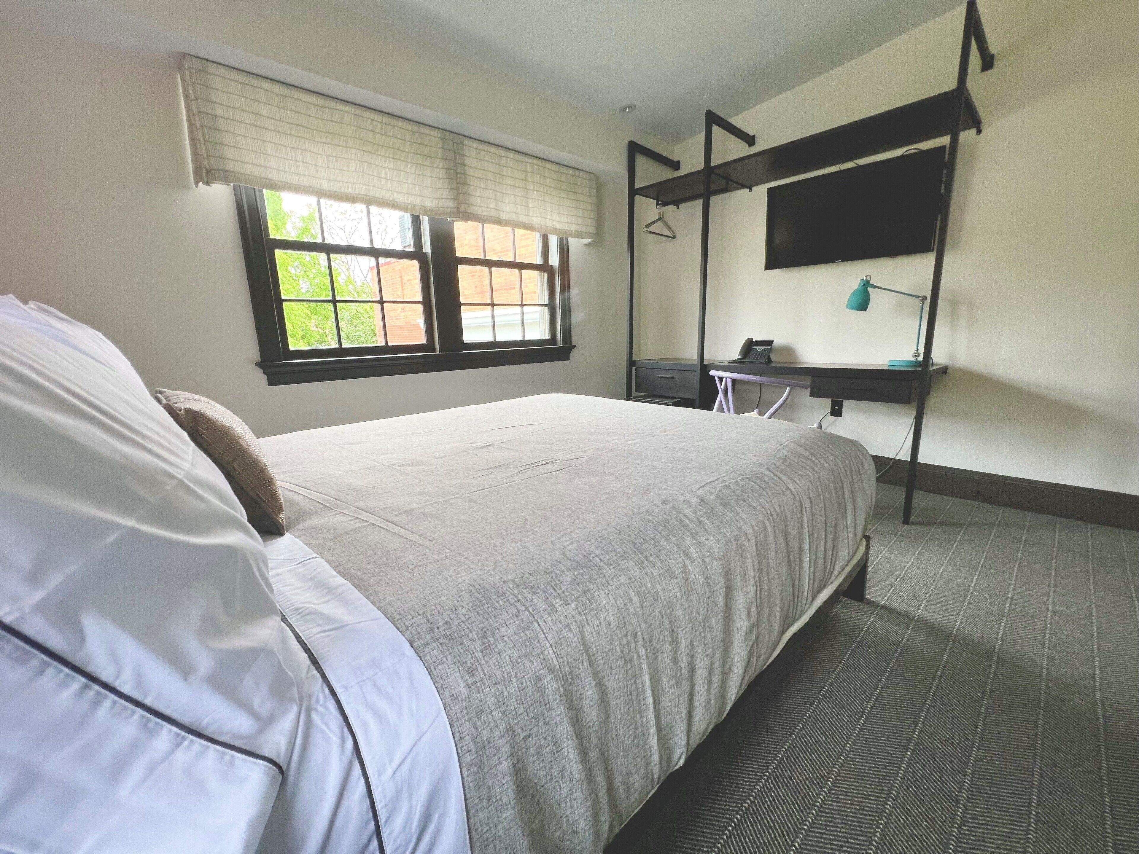 A hotel room with a bed, a desk with a chair, a TV mounted on the wall, and a window with blinds.