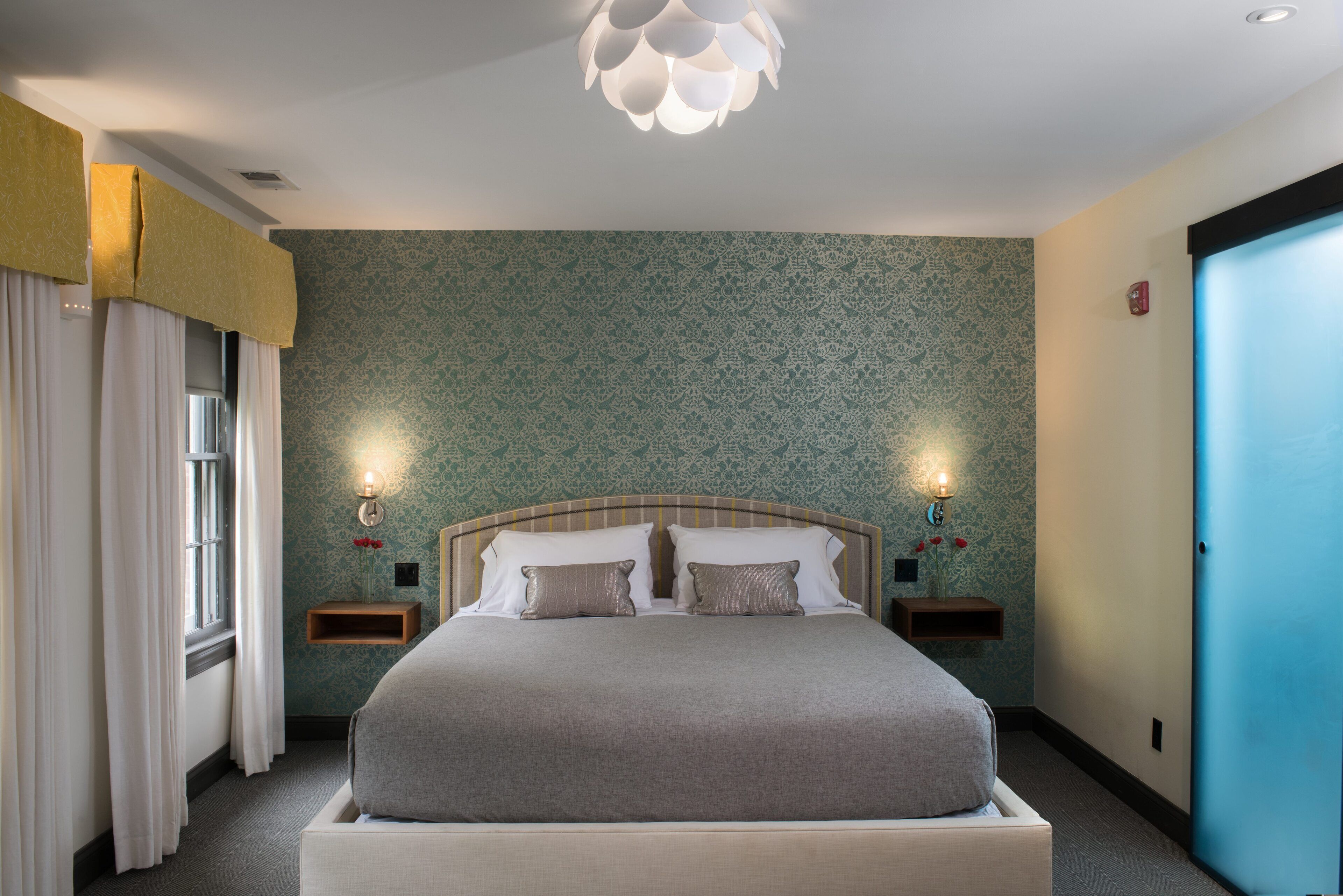 A neatly made bed with a headboard, two bedside tables with lamps, a window with curtains, and a patterned wall.
