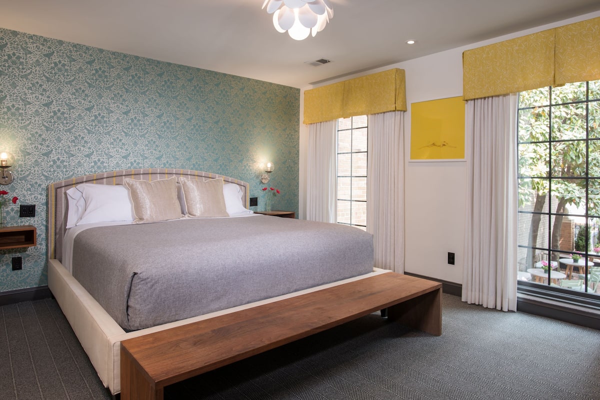 Un dormitorio con cama, mesita de noche, lámpara de pared, ventana con cortinas y vistas al exterior.