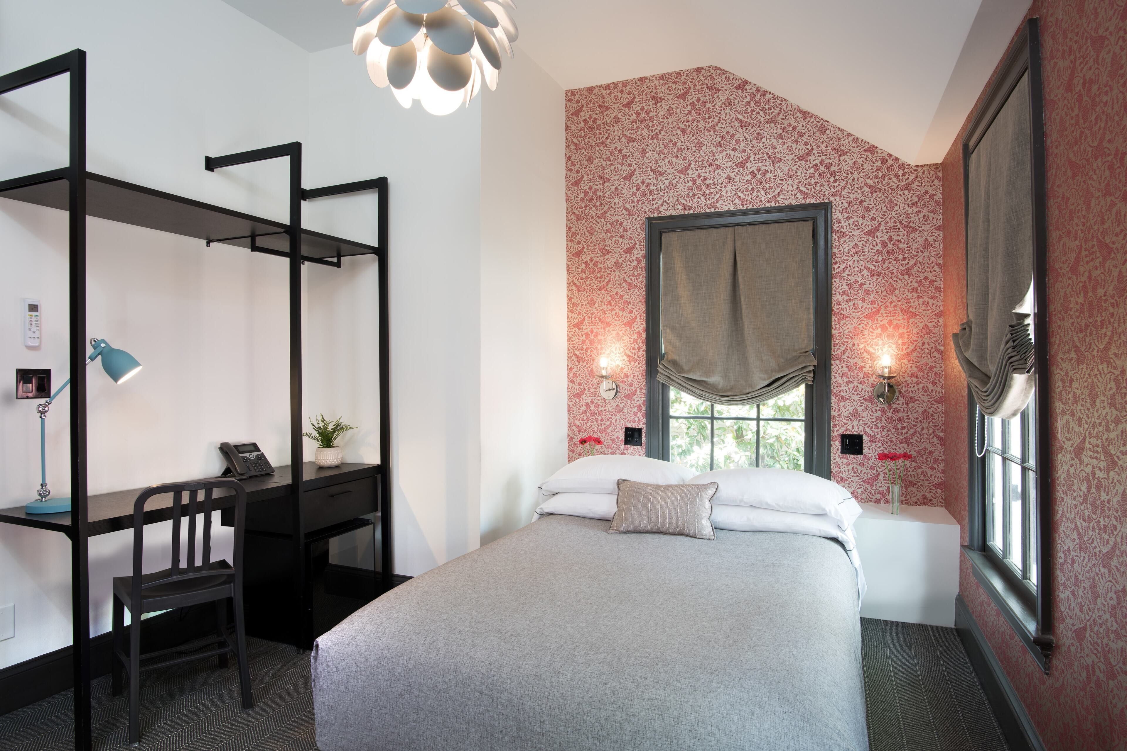 Un dormitorio con cama, escritorio con silla, escritorio de pie, ventana con cortinas y pared con papel tapiz estampado.
