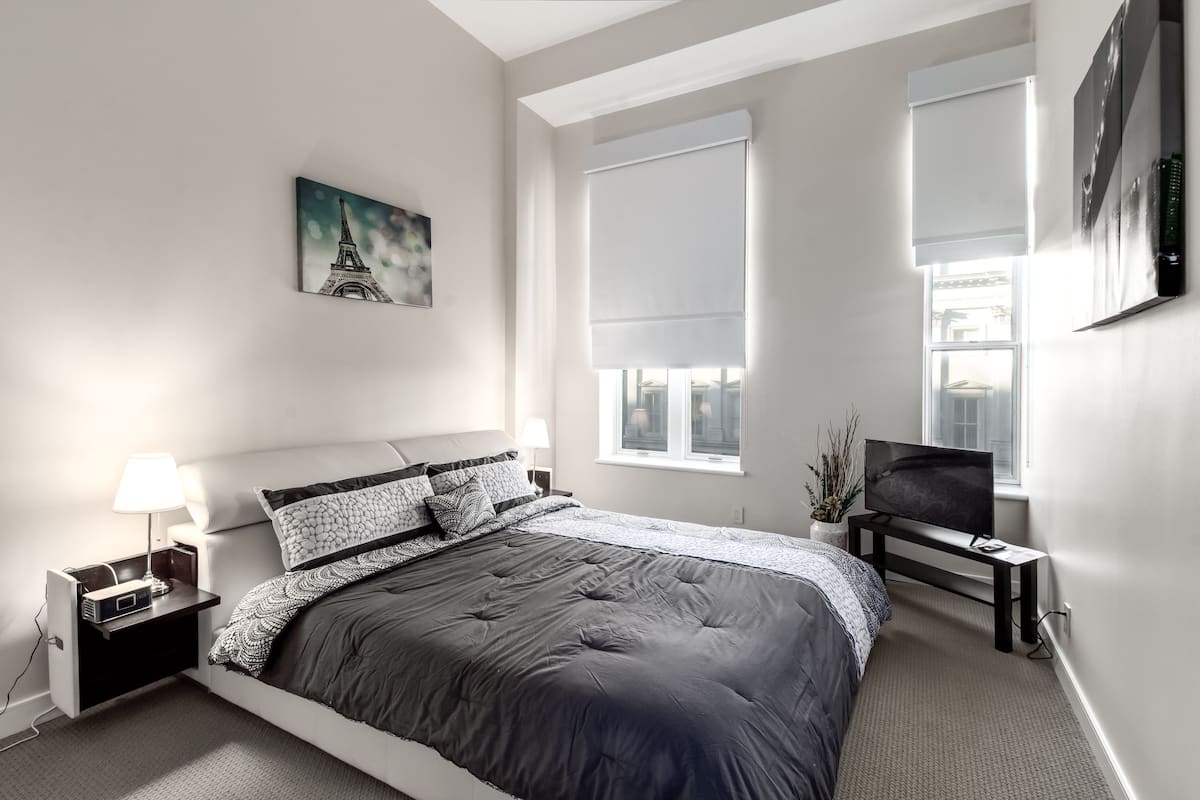 Un dormitorio moderno con una cama grande, una mesita de noche con lámpara, un televisor sobre un mueble y un cuadro de la Torre Eiffel.