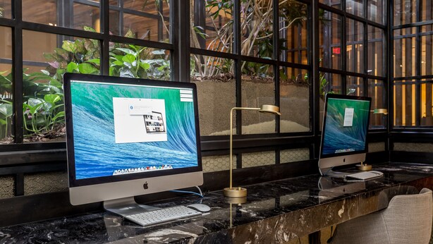 Zwei Apple iMac Computer auf einer Marmorplatte, zusammen mit einer Tastatur und einer Maus, vor einem Fenster mit Pflanzen.