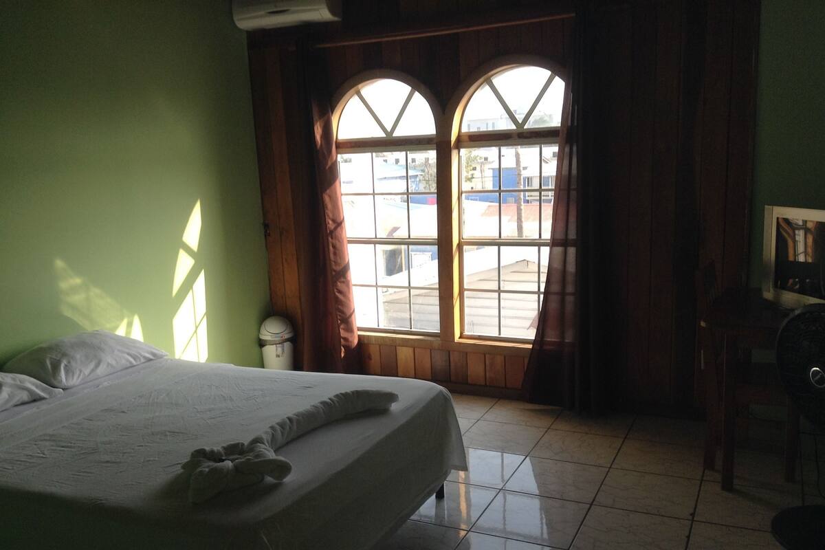 A room with a bed, a window with curtains, a chair, and a trash bin.
