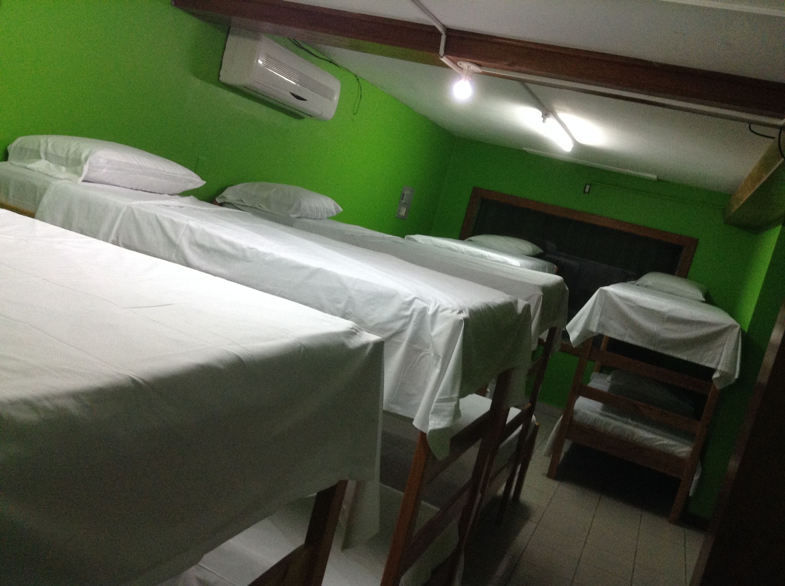 A room with three single beds, each covered with white sheets, against a green wall. There is a ceiling fan and a wall-mounted air conditioner.