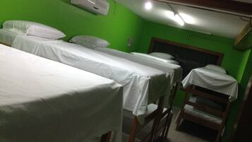 A room with three single beds, each covered with white sheets, against a green wall. There is a ceiling fan and a wall-mounted air conditioner.