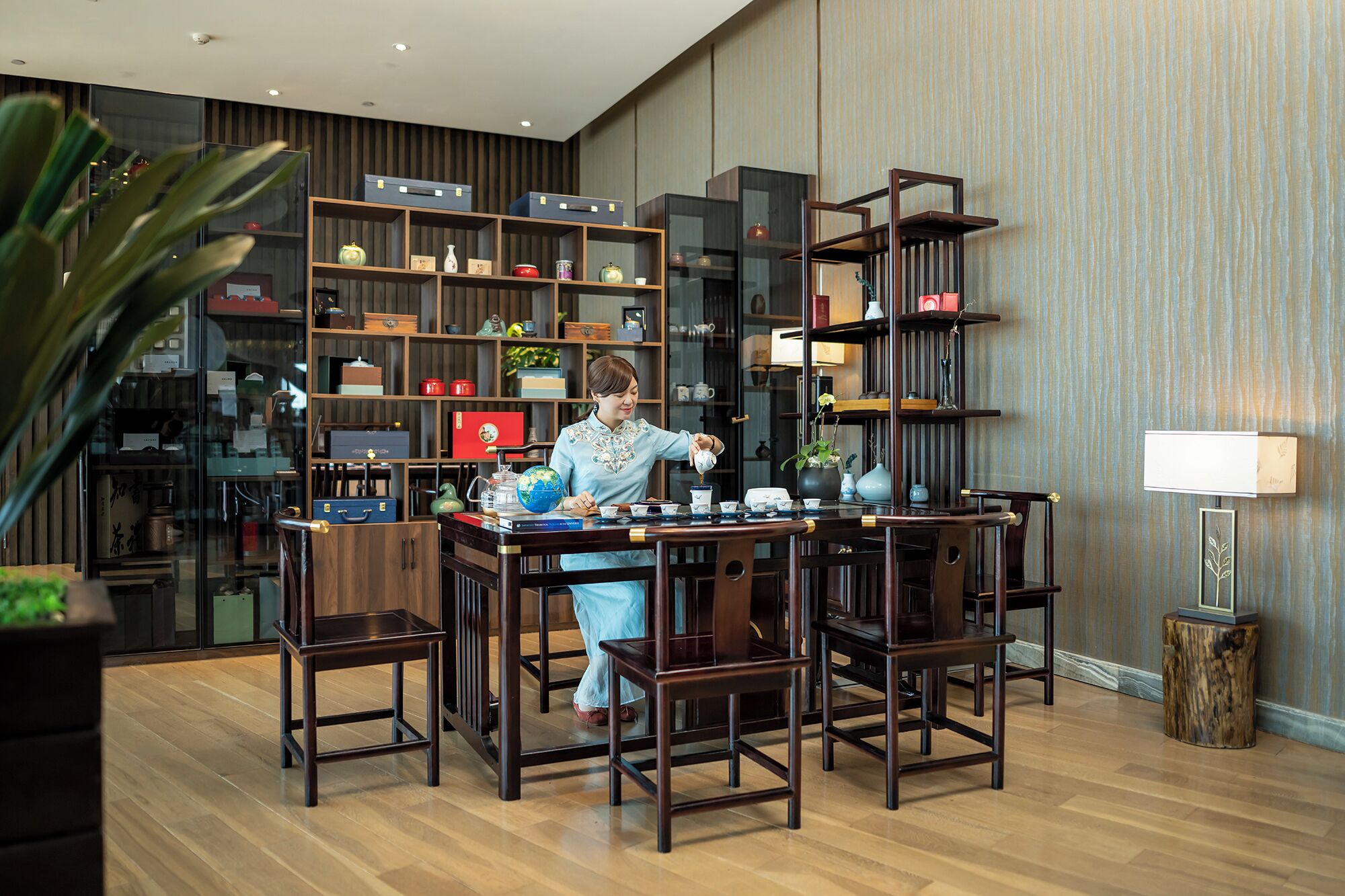 A room with a wooden floor, a table with chairs, a bookshelf with various items, and a lamp.