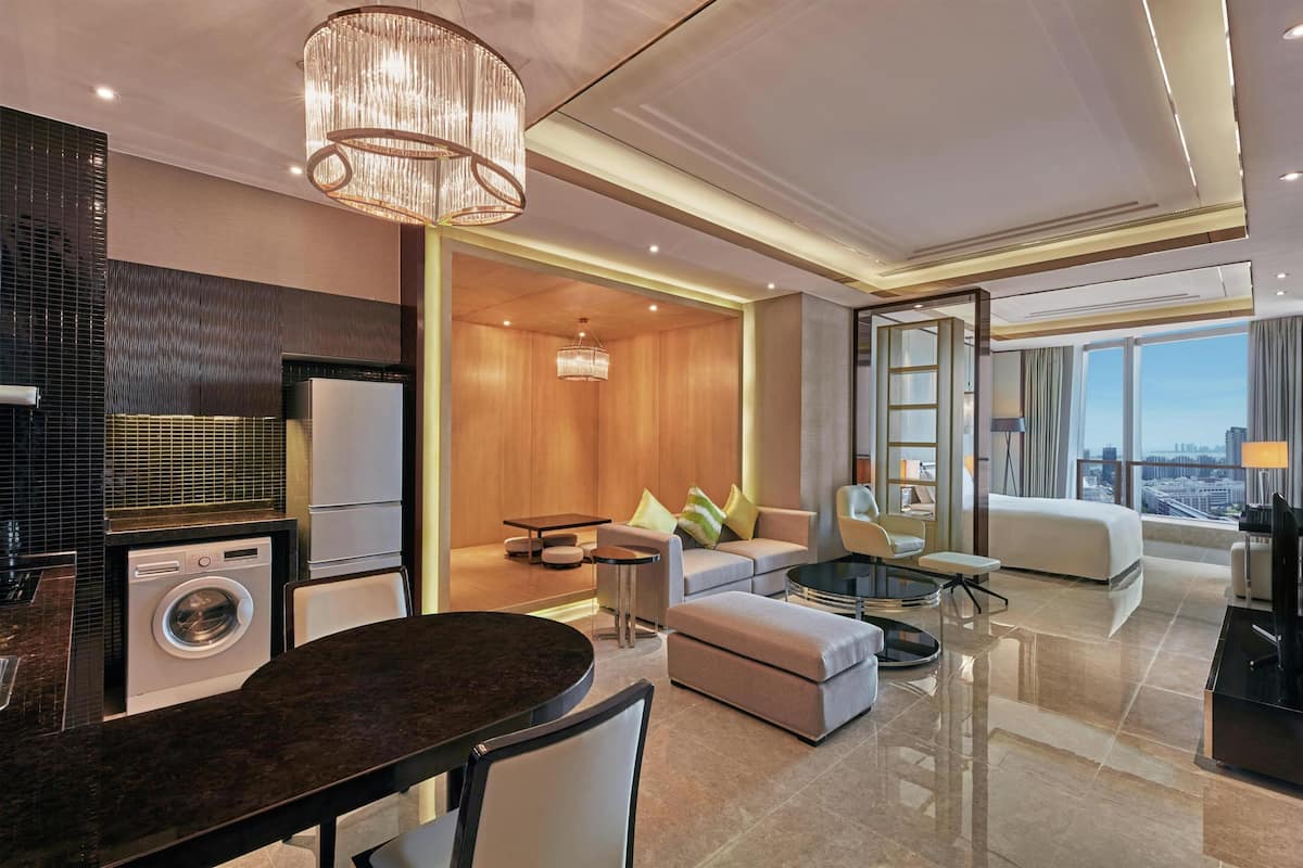 A modern hotel room with a kitchen area, a dining table, a sofa, a TV, and a large window with a city view.