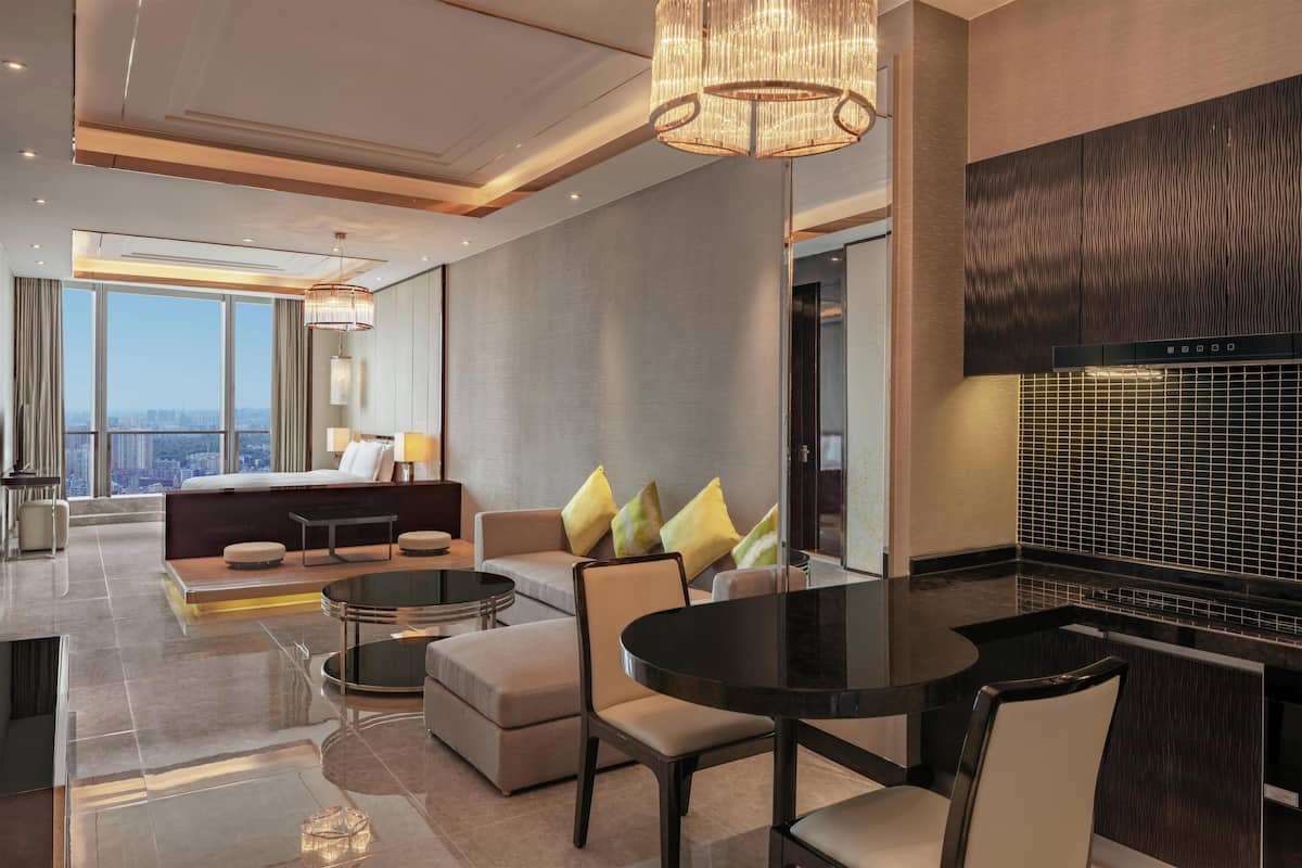 A modern hotel room with a large bed, a seating area with a sofa and chairs, a dining table, and a view of the city.