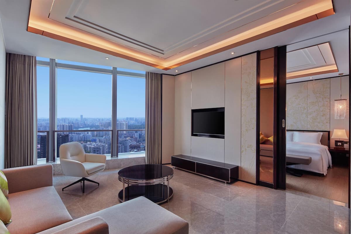 A modern hotel room with a large window, a flat-screen TV, a sofa, a coffee table, and a bed.