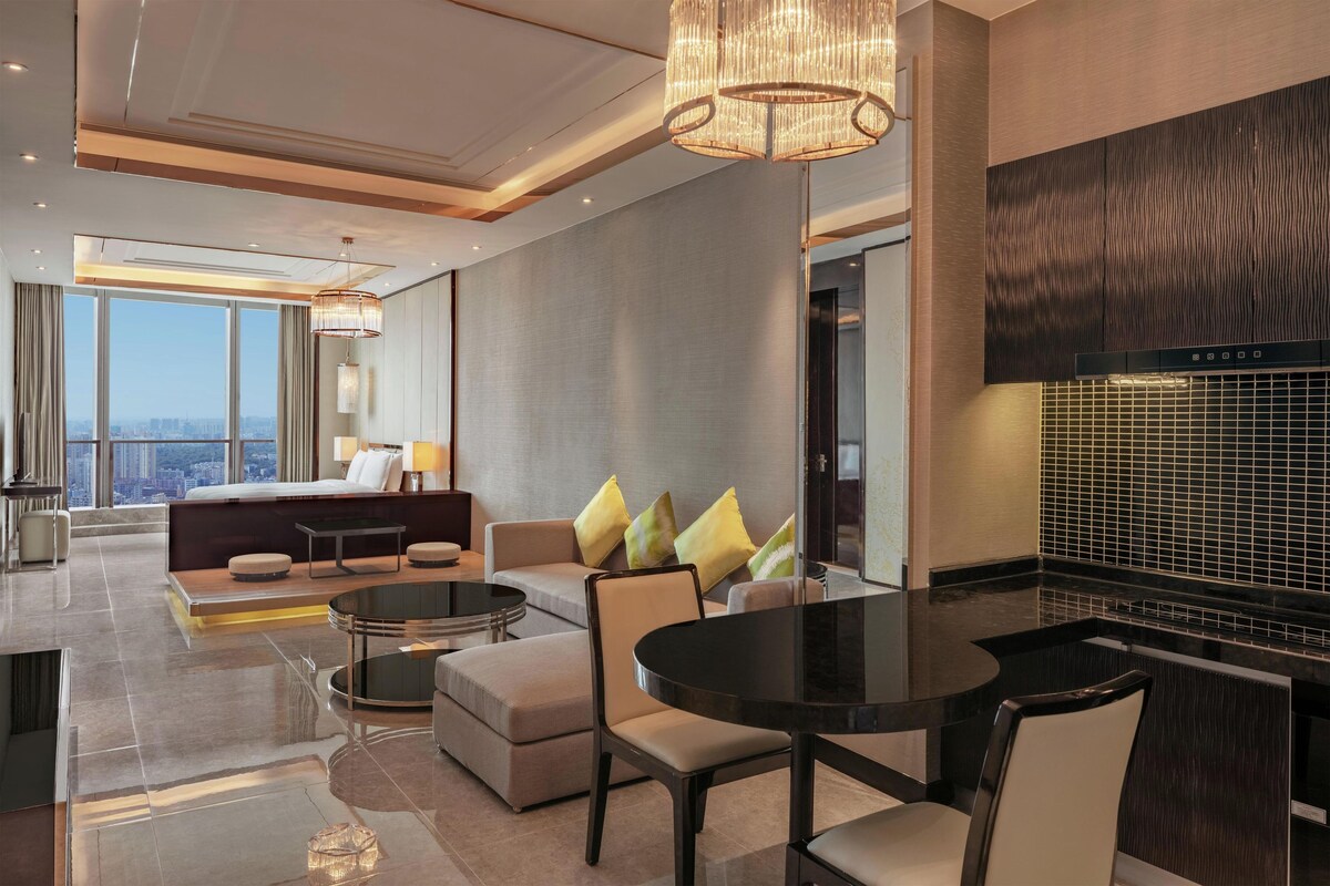 Une chambre d’hôtel moderne dotée d’un grand lit, d’un coin salon avec un canapé et des fauteuils, d’une table à manger et offrant une vue sur la ville.