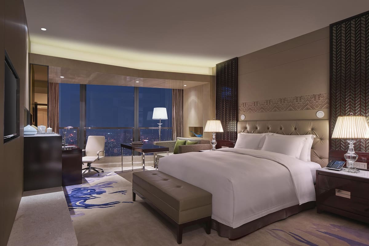 Habitación de hotel con una cama grande, un escritorio, una silla, una mesita y vista a la ciudad de noche.