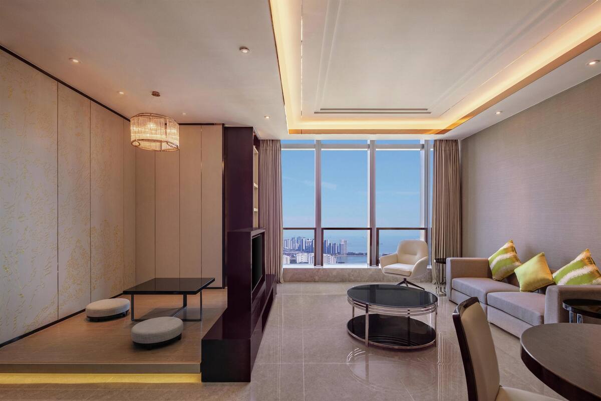 A modern living room with a sofa, ottomans, a coffee table, a TV, and a large window with a city view.