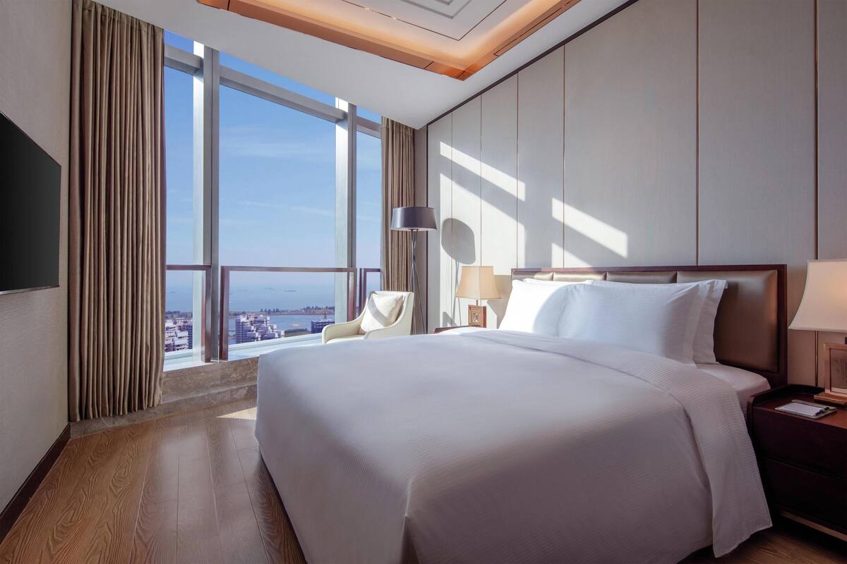 A hotel room with a large bed, a chair, a bedside table, a lamp, and a view of the city through large windows.