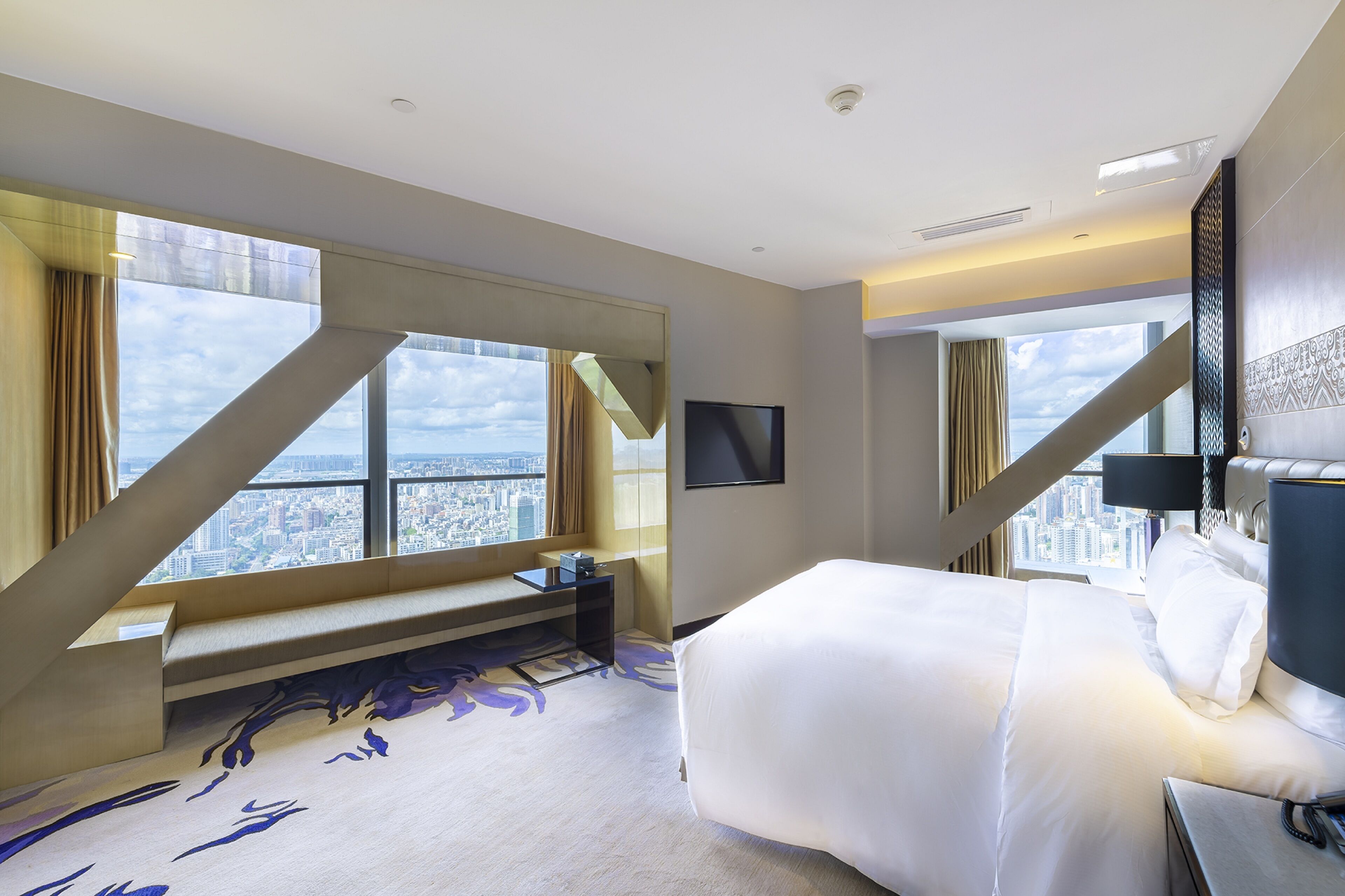 A modern hotel room with a large bed, a seating area, a flat-screen TV, and a city view.