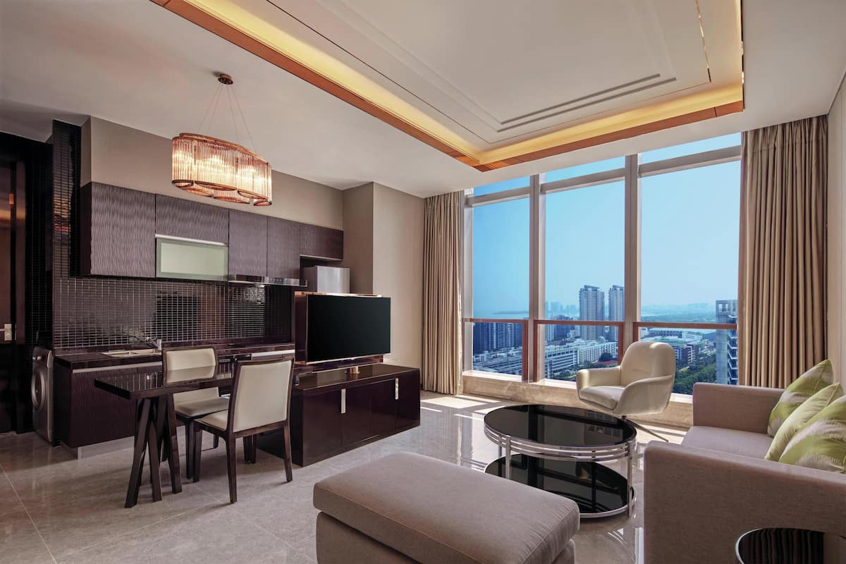 A modern hotel room with a dining area, a sofa, a TV, and a large window offering a city view.