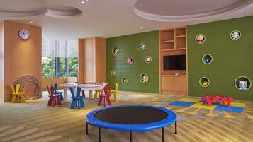 Una sala de juegos infantil con un trampolín, sillas de colores, una mesa llena de juguetes y un televisor en la pared.