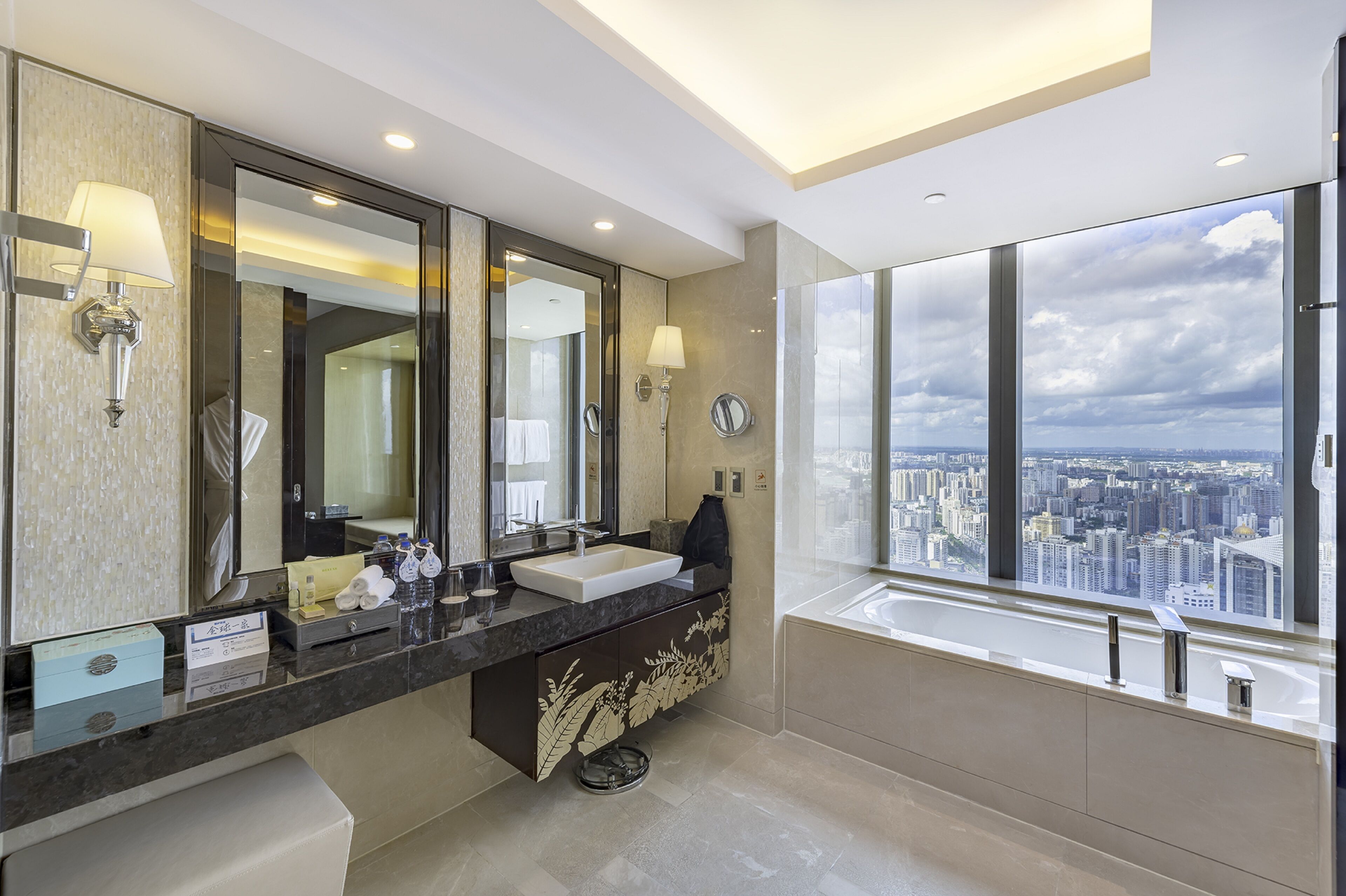 A modern bathroom with a large mirror, two sinks, and a bathtub. It features a city view through large windows and a ceiling light.
