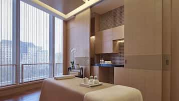 A modern spa room with a large window, a massage table, and a shelf with toiletries.