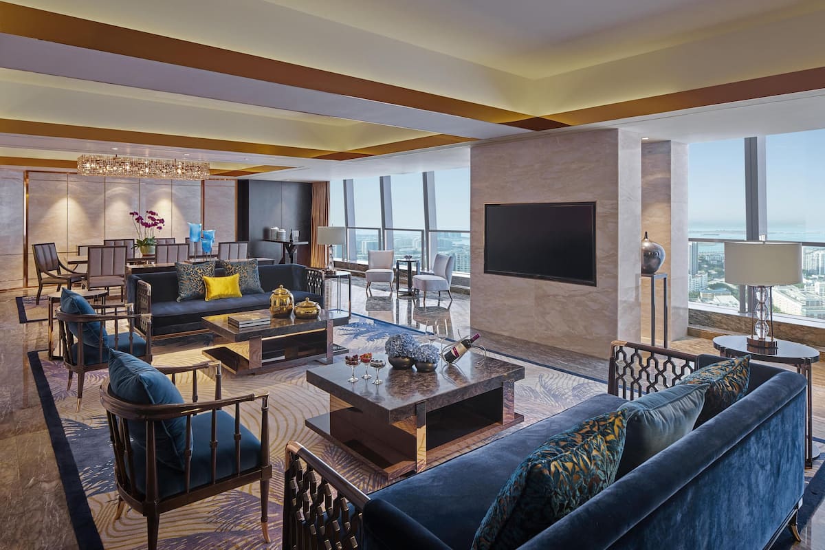 Habitación de hotel moderna con un sofá grande, zona de comedor, televisor de pantalla plana y ventanas amplias con vista a la ciudad.