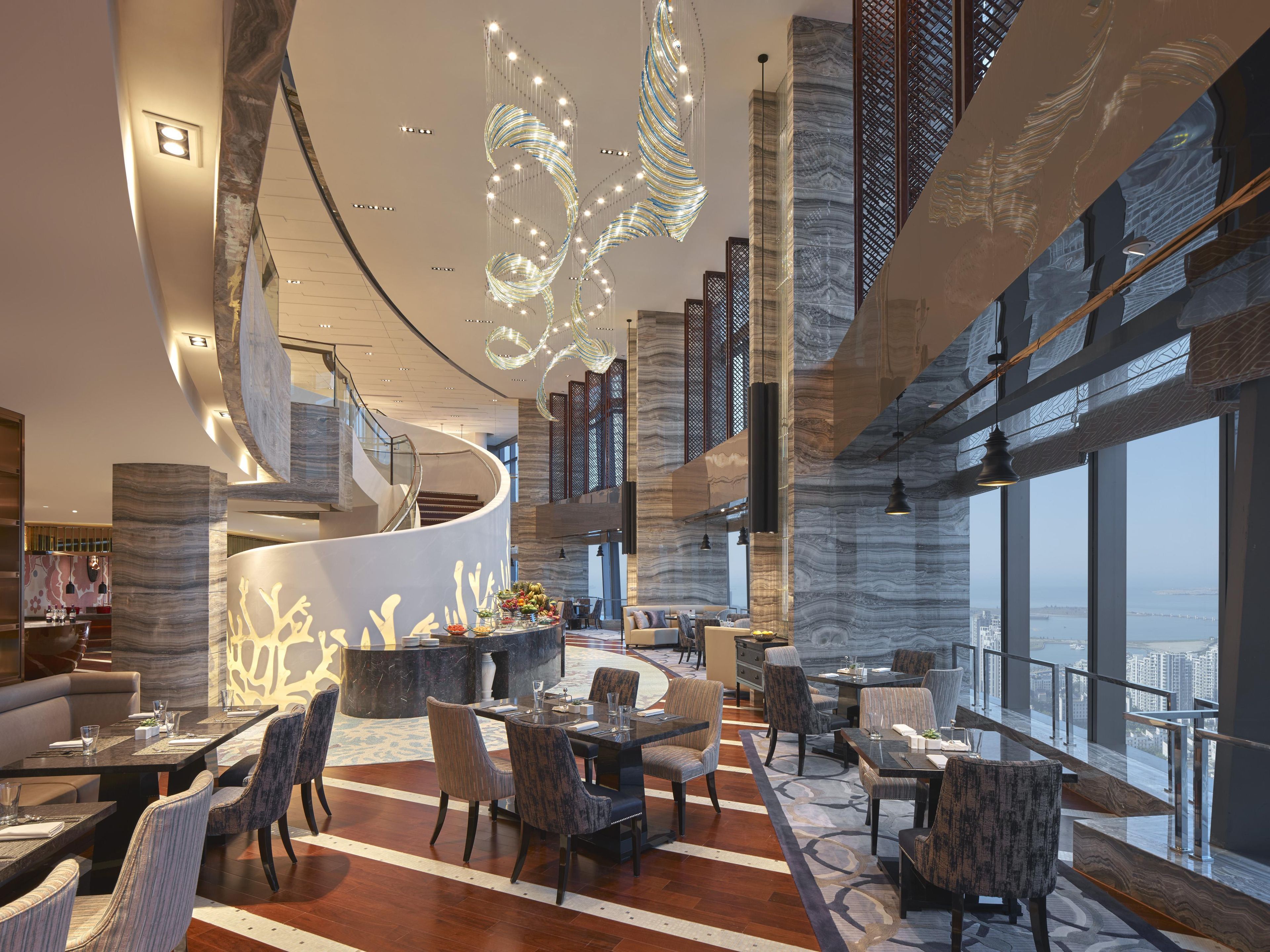 A spacious hotel lobby with a curved reception desk, multiple seating areas, and large windows offering a view of the city.