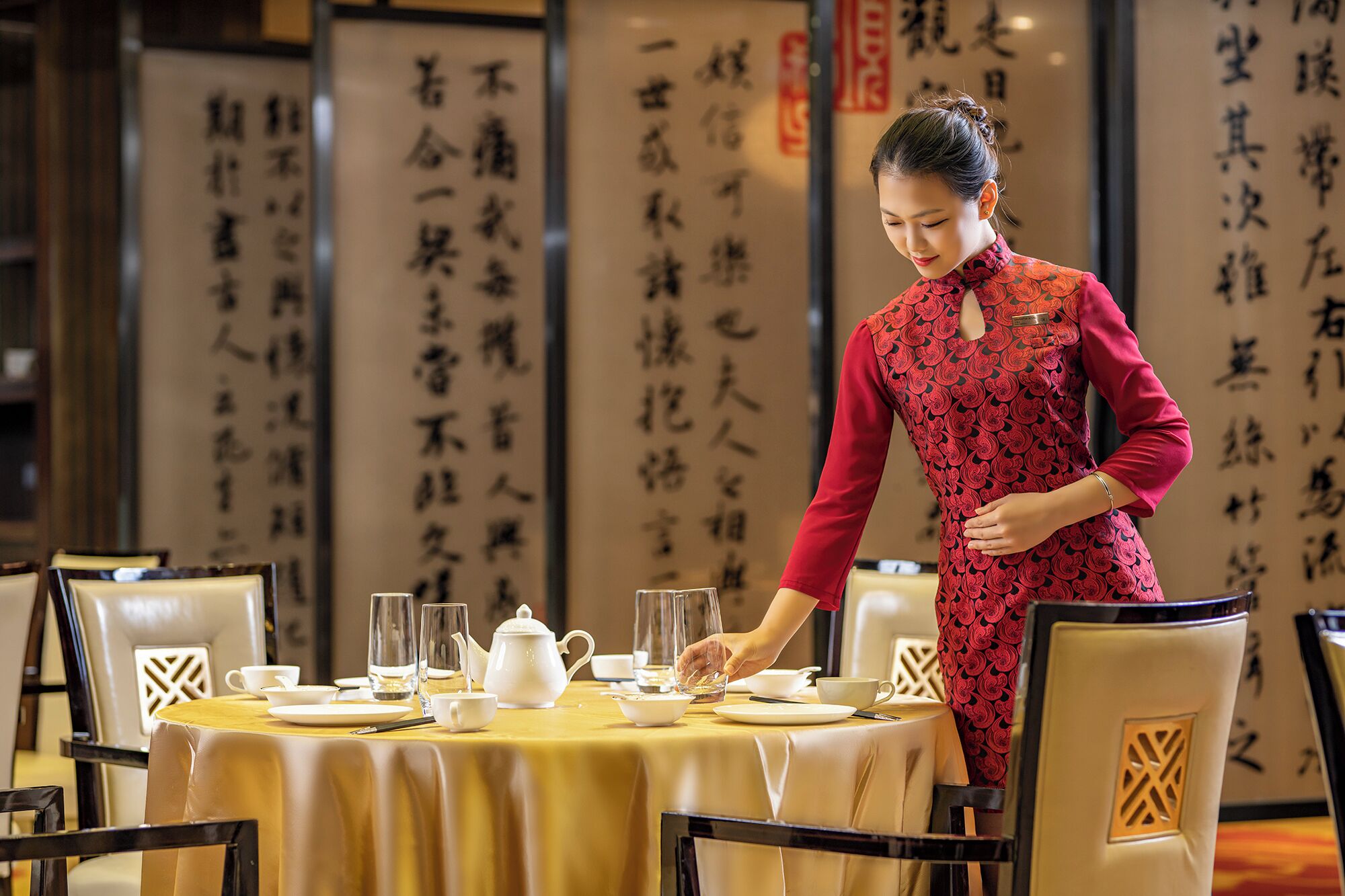 A woman in traditional attire serving at a table with tea set, surrounded by chairs and tables in a room with Chinese calligraphy on the walls.