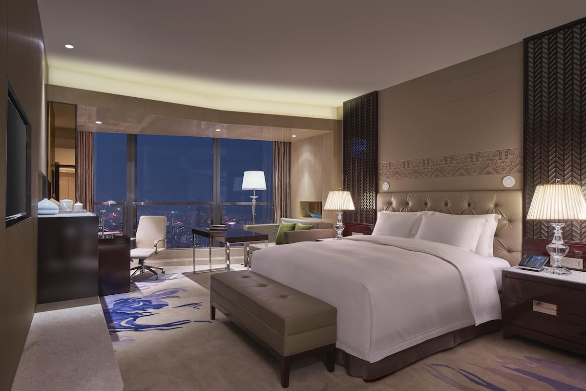 Habitación de hotel con una cama grande, un escritorio, una silla, una mesita, un banco y vistas a la ciudad de noche.