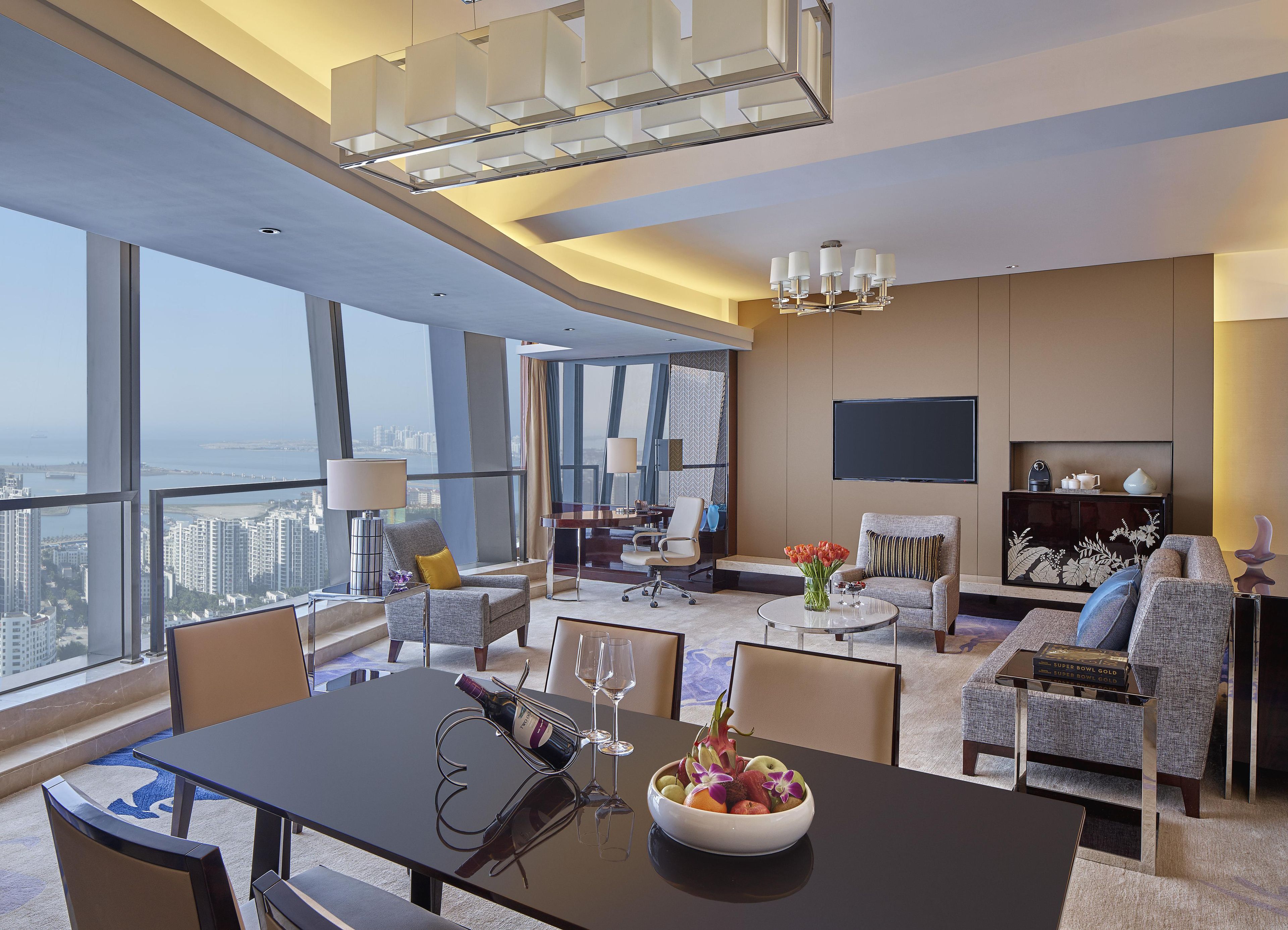 A modern hotel room with a dining area, a seating area with a sofa and armchairs, a flat-screen TV mounted on the wall, and a large window offering a city view.