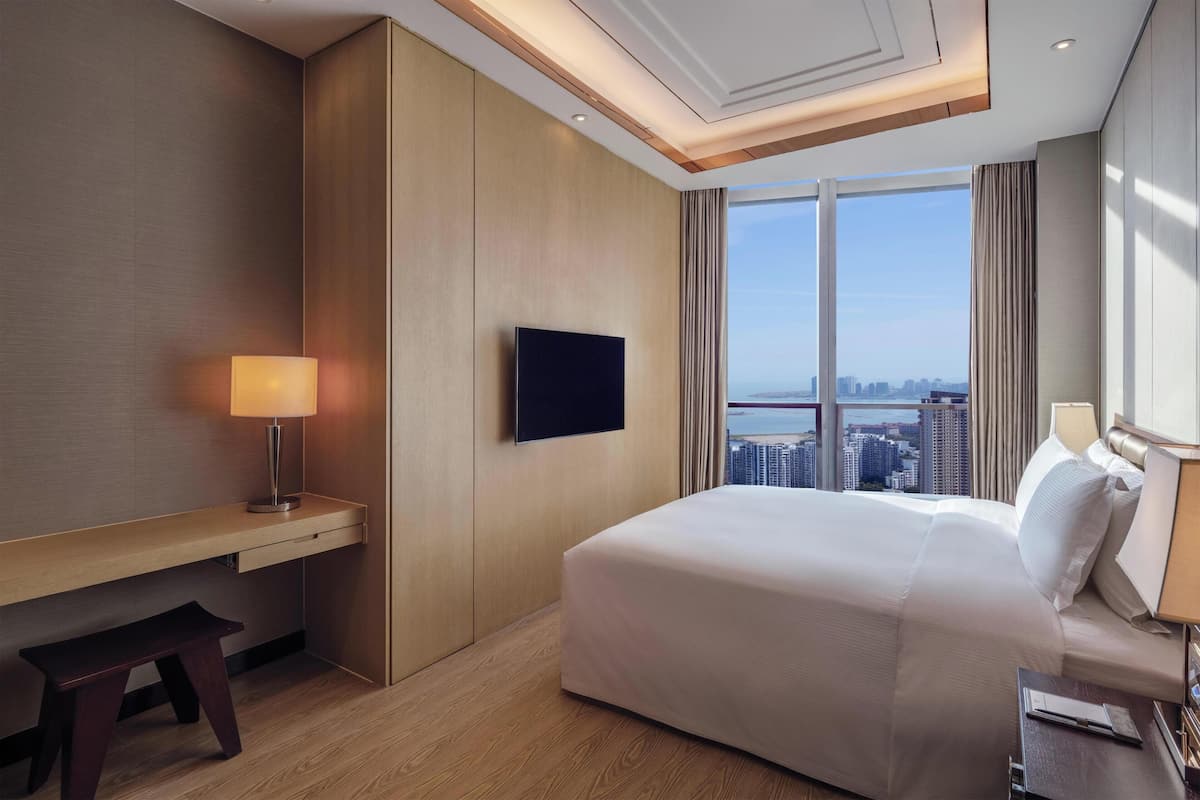 A hotel room with a bed, a TV mounted on the wall, a desk with a lamp, and a view of the city through large windows.