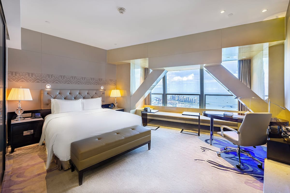 Habitación de hotel con una cama grande, un escritorio, una silla y vistas de la ciudad a través de un ventanal.