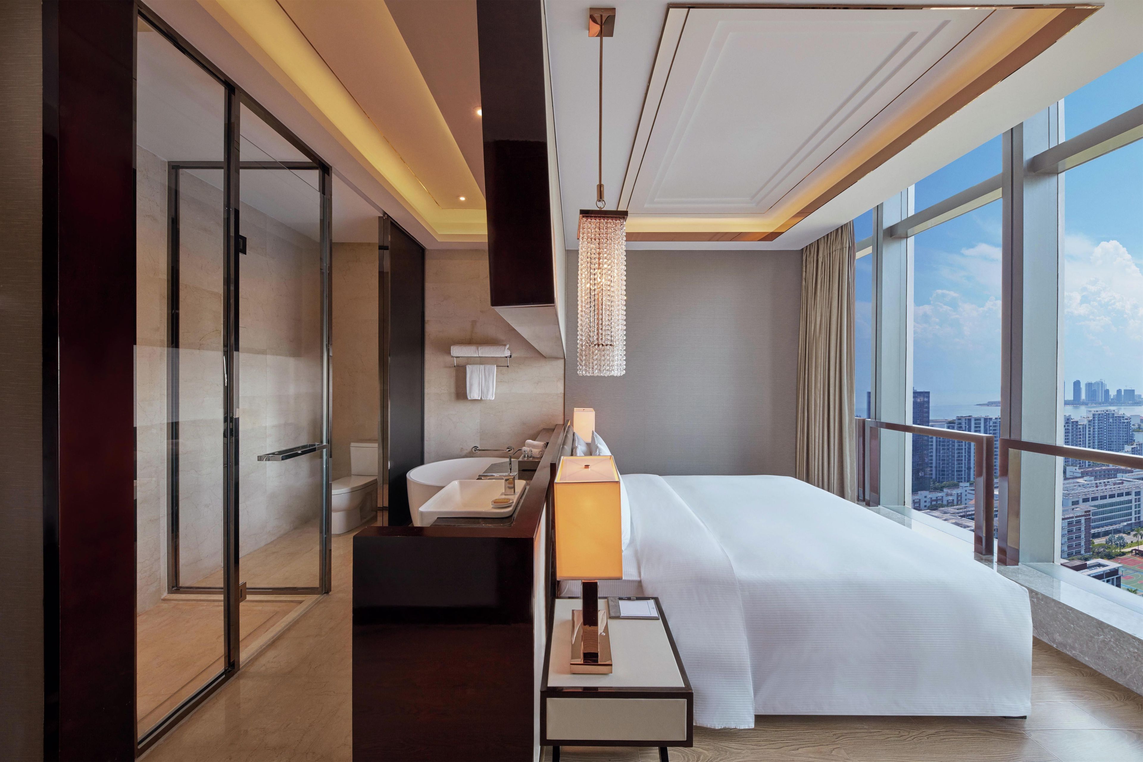 Quarto de hotel moderno com uma cama grande, banheiro com banheira, escrivaninha com luminária e vista para a cidade.