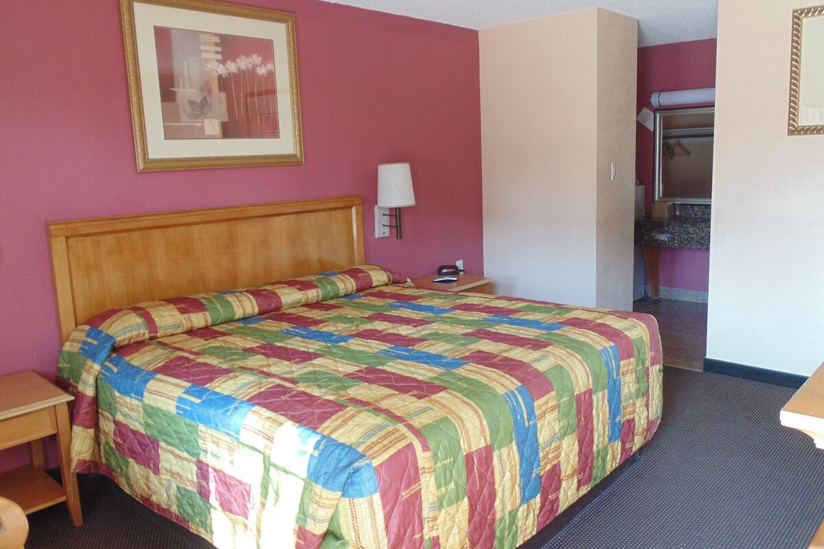 Quarto de hotel com cabeceira de madeira, colcha de patchwork colorida, mesa de cabeceira, abajur e um quadro emoldurado na parede.