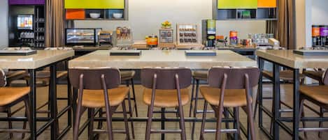 A modern cafe with a central island, bar stools, and a variety of food and drink dispensers.